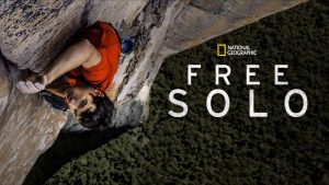 The key art image for the National Geographic documentary Free Solo. Rock climber Alex Honnold is seen climbing Yosemite’s El Capitan at left; he’s wearing a red shirt and is mid-climb, with ropes and pulleys attached to him. At right, the precarious drop down to the ground can be seen behind the National Geographic logo and the title of the film. 