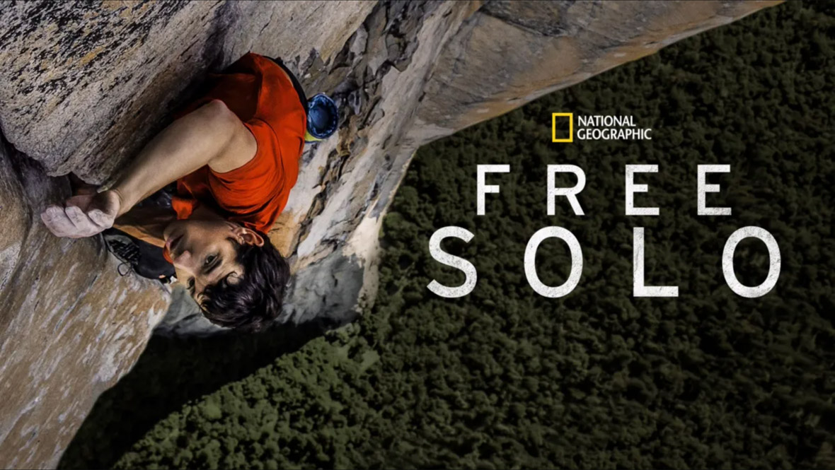 The key art image for the National Geographic documentary Free Solo. Rock climber Alex Honnold is seen climbing Yosemite’s El Capitan at left; he’s wearing a red shirt and is mid-climb, with ropes and pulleys attached to him. At right, the precarious drop down to the ground can be seen behind the National Geographic logo and the title of the film. 