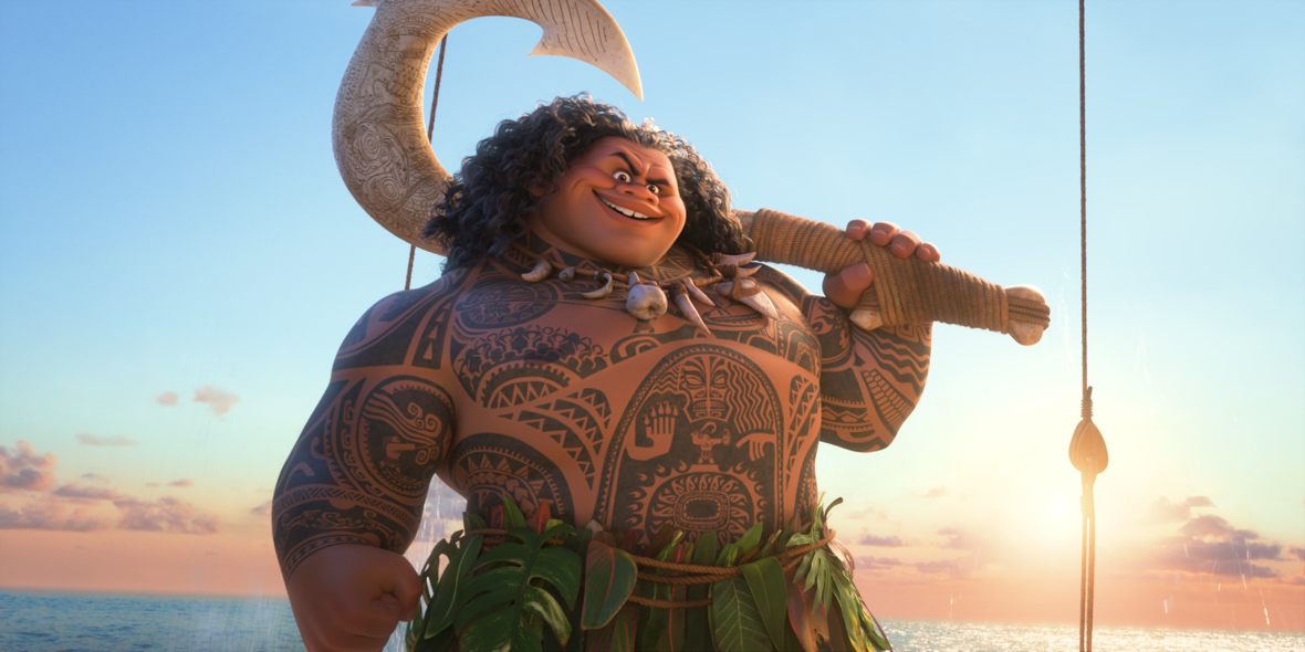 In an image from Walt Disney Animation Studios’ Moana 2, Maui (voiced by Dwayne Johnson) is standing looking at the camera, holding his hook over one shoulder. The sun on the horizon, and the ocean, are seen behind him.