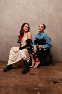 A promotional image of Moana 2 songwriters Abigail Barlow (left) and Emily Bear (right). Barlow has long, dark red hair and is wearing a strapless white gown, black gloves, and black boots. Bear has pulled-back dark hair and is wearing a black and denim top with a black-and-white polka-dot skirt, and black and white heels. They are sitting on a trunk and looking at the camera.