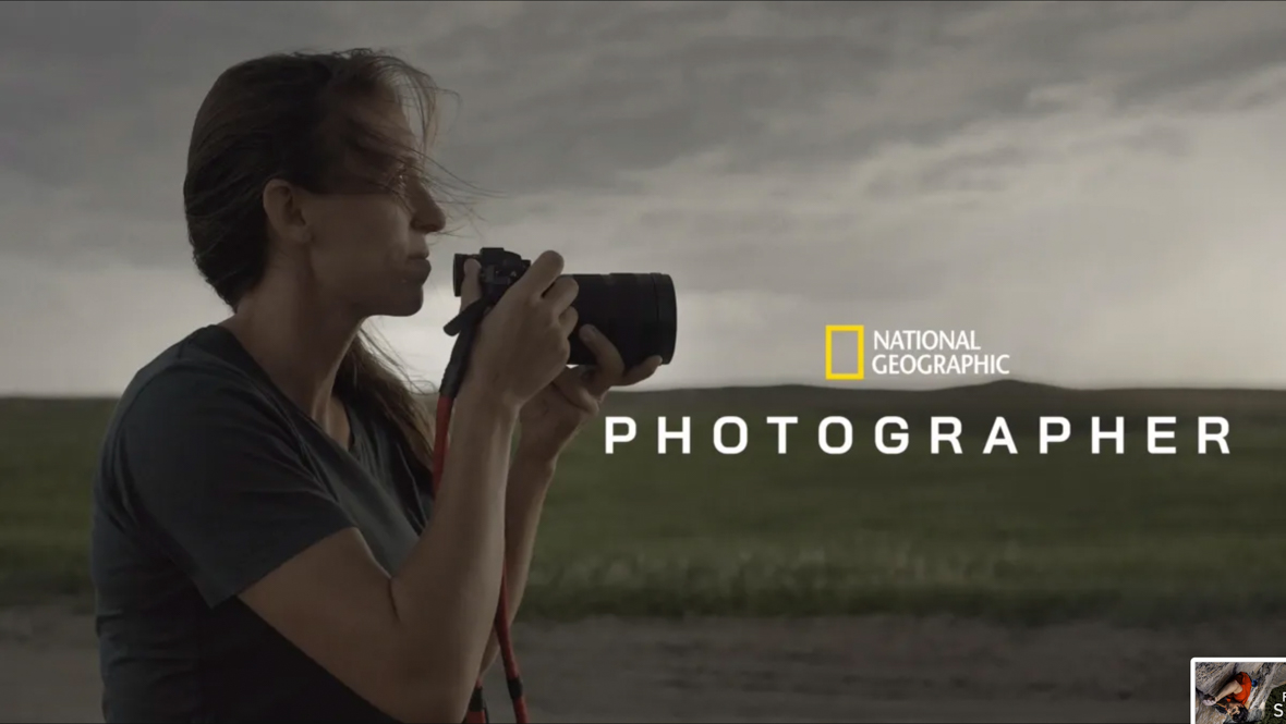 A key art image for the National Geographic series Photographer. At left, a woman is holding a camera; she’s wearing a gray shirt and her hair, partially in a ponytail, is windswept. Behind her is a field. To the right, set against the field, is the National Geographic logo and the title of the series. 