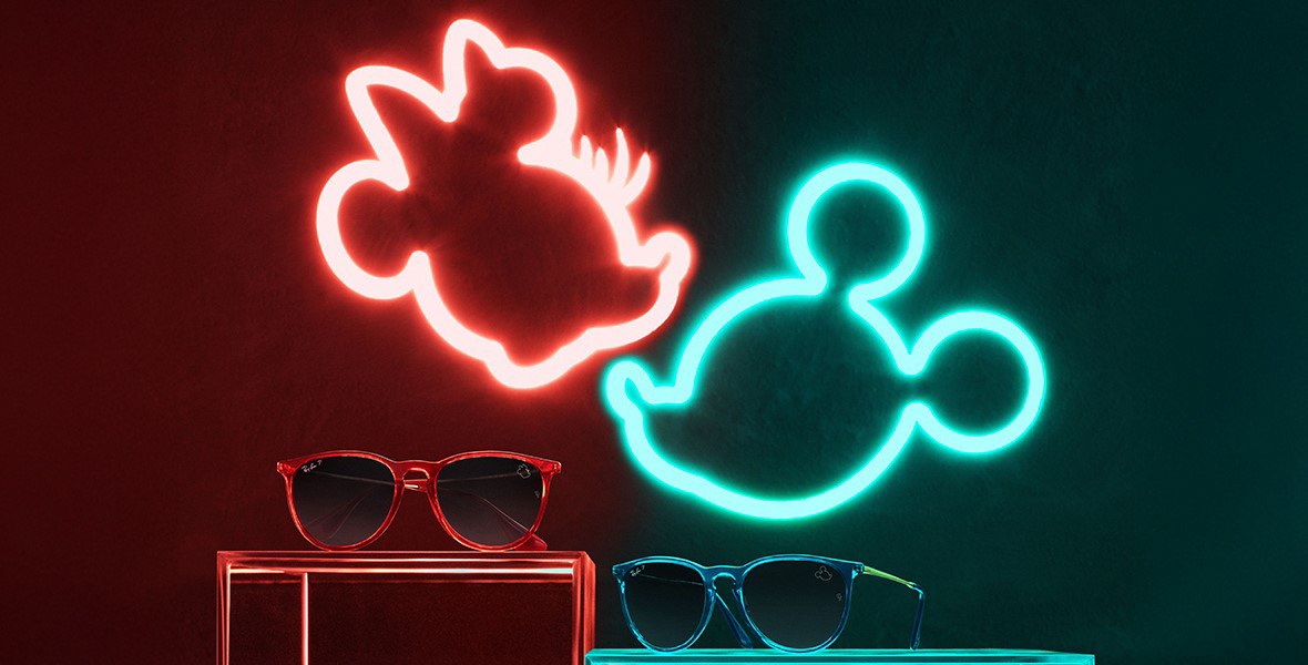 (from left to right) A red pair of Ray-Ban sunglasses sits atop a red table and green pair of Ray-Ban’s sit on a green table. The red has a neon-lit icon of Minnie Mouse and atop the green a neon-green icon of Mickey Mouse from Sunglass Hut’s Mickey Mouse Collection.