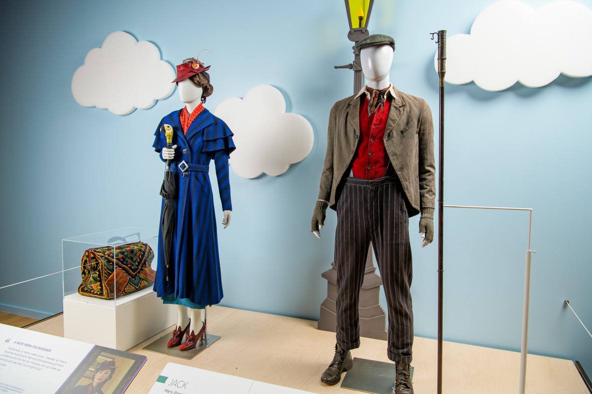 The costumes for Mary Poppins and Jack from Mary Poppins Returns displayed in the exhibit against a background of a blue sky and clouds in the Heroes & Villains: The Art of the Disney Costume exhibit.