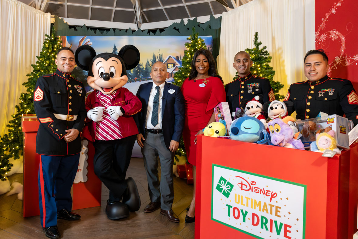 In an image from the Mickey Around the World Toy Drive Tour, Mickey Mouse poses with Disney cast members and U.S. Marines at the NYC Disney store, with a box filled with Disney plush toys that reads “Disney Ultimate Toy Drive” in the bottom right of the photo. The background features lit Christmas trees.