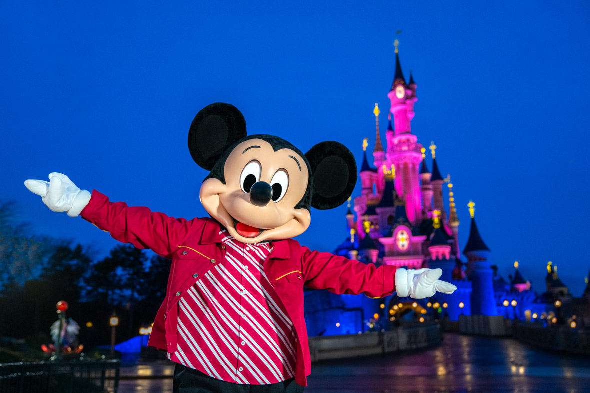 In an image from the Mickey Around the World Toy Drive Tour, Mickey Mouse poses in front of the Sleeping Beauty Castle at Disneyland Paris at night.