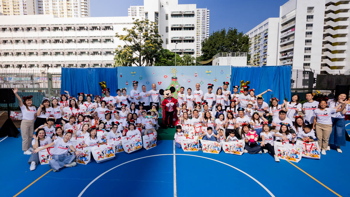 In an image from the Mickey Around the World Toy Drive Tour, Mickey Mouse poses with students, teachers, and Disney VoluntEARS on an outdoor basketball court. The background features tall white buildings.