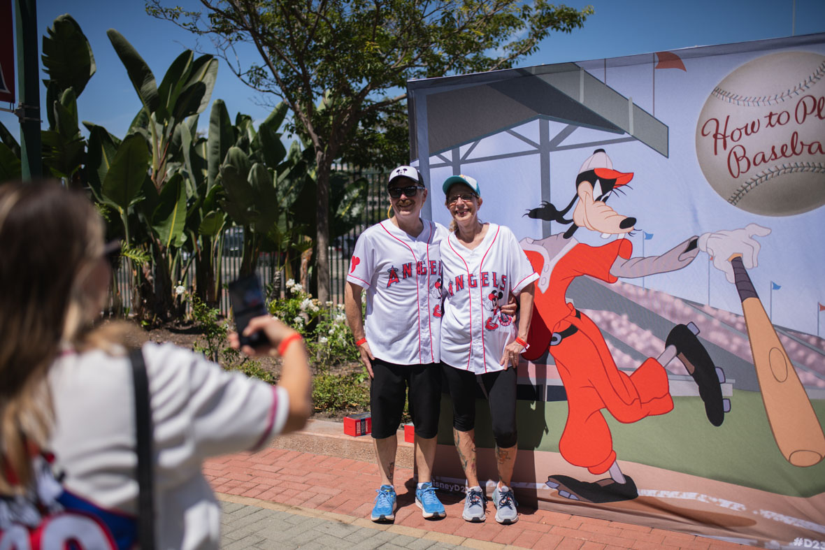 An image from D23 Day at Angel Stadium shows two fans posing in front of a Goofy photo op. Goofy is dressed in a red baseball uniform, holding a bat and a baseball featuring “How to Play Baseball.” A third guest takes a photo of the pair, who are wearing white baseball shirts that read “ANGELS.”