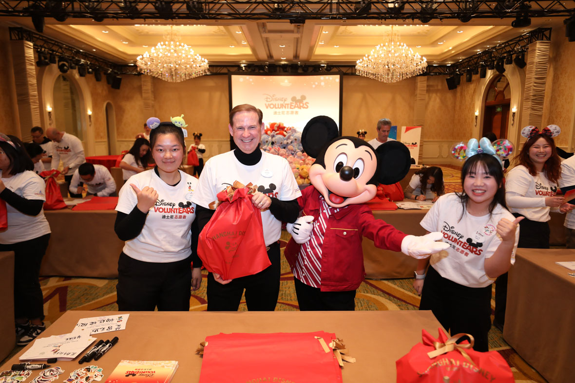 In an image from the Mickey Around the World Toy Drive Tour, Mickey Mouse poses with Disney VoluntEARS as they wrap up presents. The background features an interior space where other VoluntEARS are working.