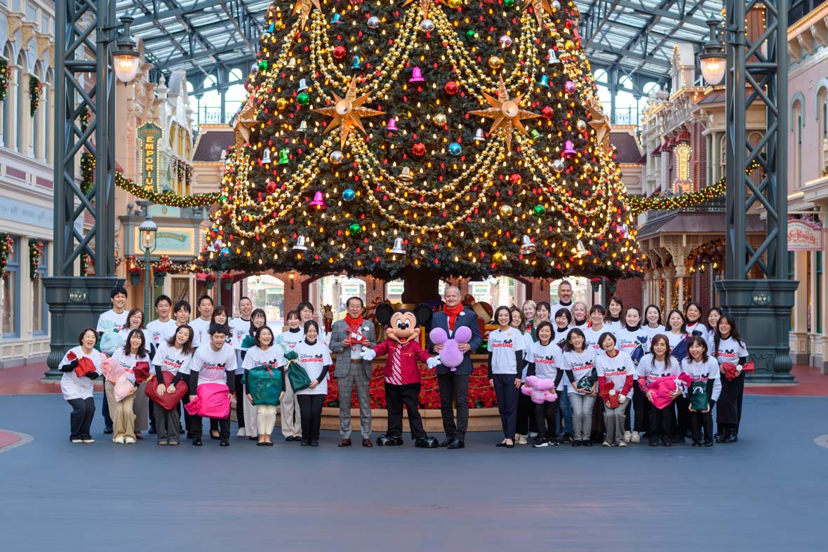 In an image from the Mickey Around the World Toy Drive Tour, Mickey Mouse poses with Tokyo Disney Resort Ambassadors and cast members in front of a lit Christmas tree.