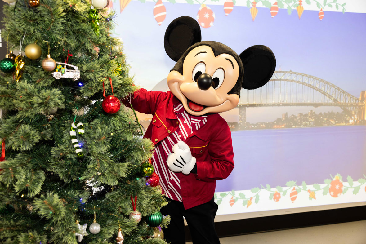 In an image from the Mickey Around the World Toy Drive Tour, Mickey Mouse poses next to a decorated Christmas tree. The background features an image of the Sydney Harbour Bridge.