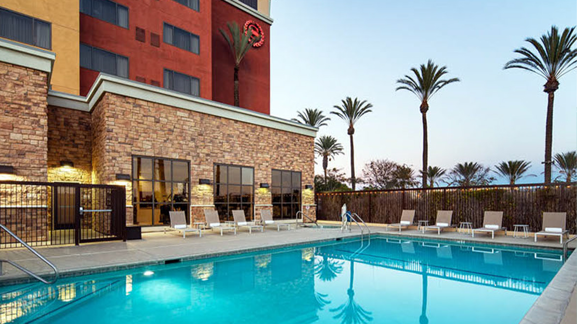 The exterior of the Sheraton Garden Grove in Anaheim, CA. Various lounge chairs with rolled towels are set up on the concrete alongside the hotel’s pool. The pool has two sets of steps under the water with a railing to hold onto. Behind the pool (and to the right of the hotel building) are rows of palm trees against a blue sky; they reflect in the pool’s water.