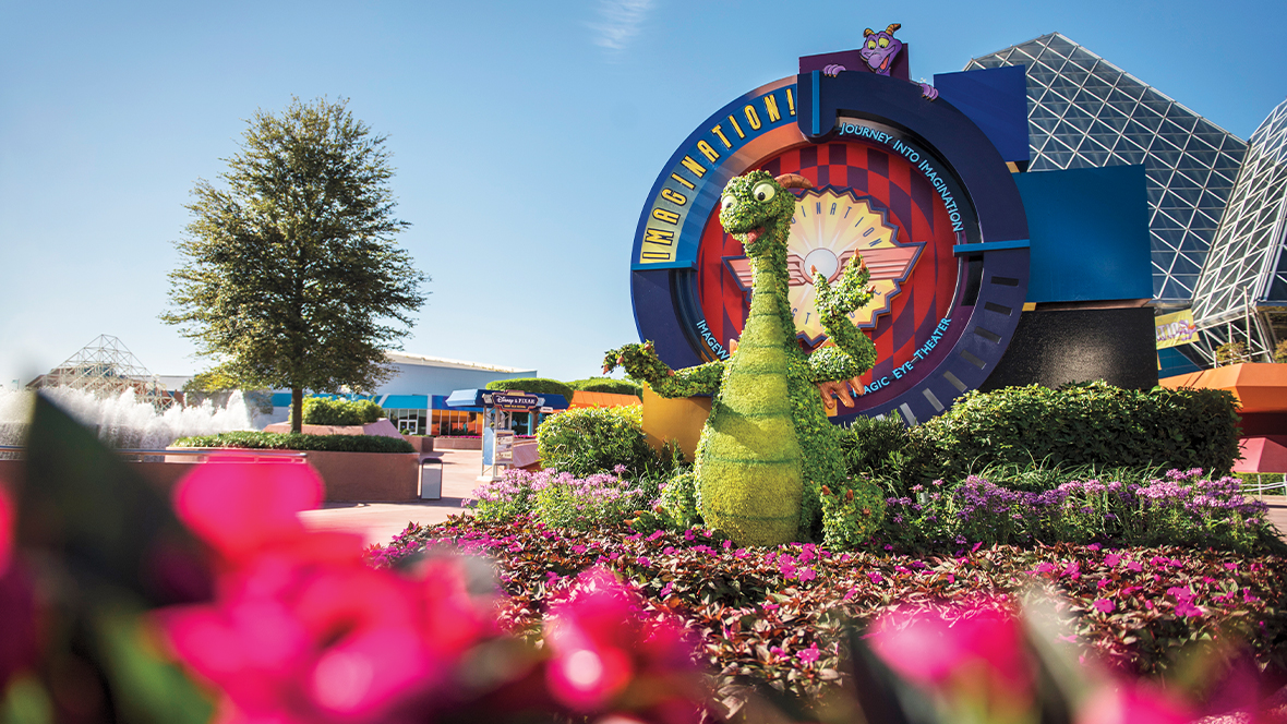 An image from the EPCOT International Flower & Garden Festival at Walt Disney World Resort features Figment, assembled with green leaves. A sign behind reads “IMAGINATION! JOURNEY INTO IMAGINATION,” with an animated Figment on the top of the sign, looking towards the statue of Figment. The background features a building with solar roof panels, a tree, and a clear blue sky.   