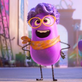 In an image from Dream Productions, Paula (voiced by Paula Pell), a purple, anthropomorphic, jellybean-like director, sports a purple wig, orange glasses, and an orange necktie. Beside her is a blue, jellybean-like dog with a purple nose, ears, and tail, and a pink tongue. The backdrop showcases the vibrant film studio Dream Productions.