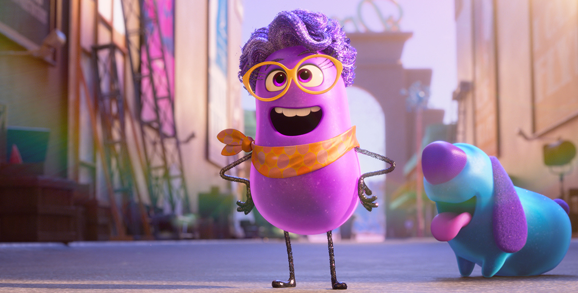 In an image from Dream Productions, Paula (voiced by Paula Pell), a purple, anthropomorphic, jellybean-like director, sports a purple wig, orange glasses, and an orange necktie. Beside her is a blue, jellybean-like dog with a purple nose, ears, and tail, and a pink tongue. The backdrop showcases the vibrant film studio Dream Productions.