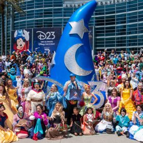 In an image from D23: The Ultimate Disney Fan Event, fans cosplaying as Disney characters gather in front of the Anaheim Convention Center around a huge, sparkling Mickey Mouse Sorcerer Hat. They look at the camera, smiling and waving. Palm trees are in the background.