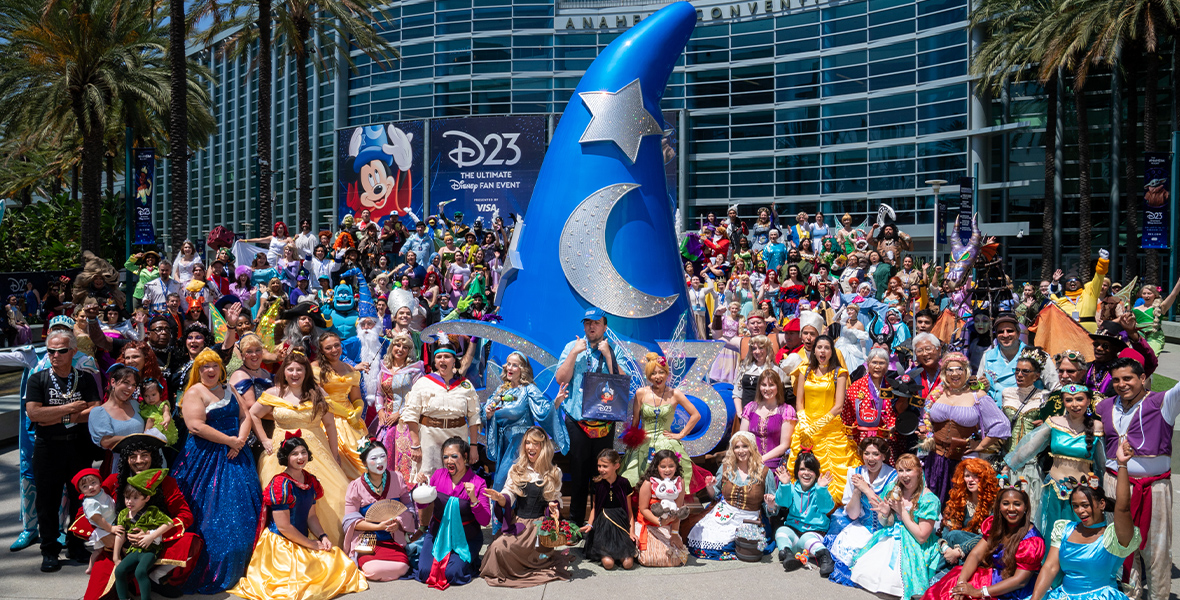 In an image from D23: The Ultimate Disney Fan Event, fans cosplaying as Disney characters gather in front of the Anaheim Convention Center around a huge, sparkling Mickey Mouse Sorcerer Hat. They look at the camera, smiling and waving. Palm trees are in the background.