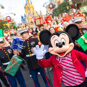 Mickey Mouse waves to the camera at Disneyland’s Main Street, U.S.A., during the Mickey Around the World Toy Drive Tour. Behind him are Disney VoluntEARS and U.S. Marines, holding wrapped gifts and smiling. The background features the Disneyland’s Sleeping Beauty Castle and shops with wreaths.