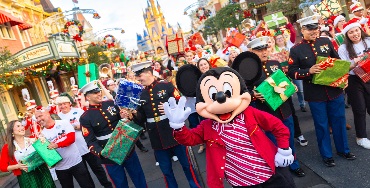 Mickey Mouse waves to the camera at Disneyland’s Main Street, U.S.A., during the Mickey Around the World Toy Drive Tour. Behind him are Disney VoluntEARS and U.S. Marines, holding wrapped gifts and smiling. The background features the Disneyland’s Sleeping Beauty Castle and shops with wreaths.