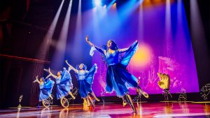 Four performers in bright blue dresses balance on unicycles as they dance in synchronization on a stage. The stage is lit by multiple overhead lights spotlighting the performers. The backdrop is multicolored with various shades of blue and purple that all blend into one another. Behind the unicycle dancers is a performer who is wearing the costume of a giant hand from the waste up.