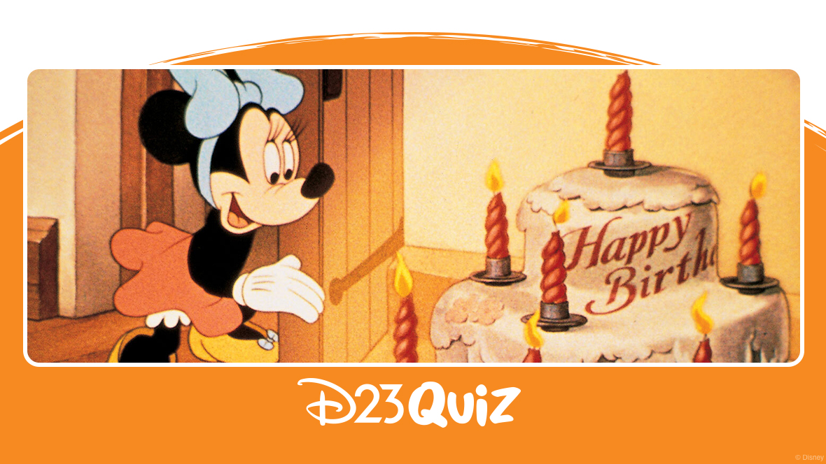 In an image from Mickey’s Birthday Party, Minnie Mouse (voiced by Thelma Boardman) wears a light blue bow, a red skirt, and yellow shoes. She stands behind an open door, looking at a tiered cake with candles that read “Happy Birthday.” The background features the interior of a room with wooden beams on the floor.