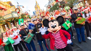 Mickey Mouse waves to the camera at Disneyland&rsquo;s Main Street, U.S.A., during the Mickey Around the World Toy Drive Tour. Behind him are Disney VoluntEARS and U.S. Marines, holding wrapped gifts and smiling. The background features the Disneyland&rsquo;s Sleeping Beauty Castle and shops with wreaths.
