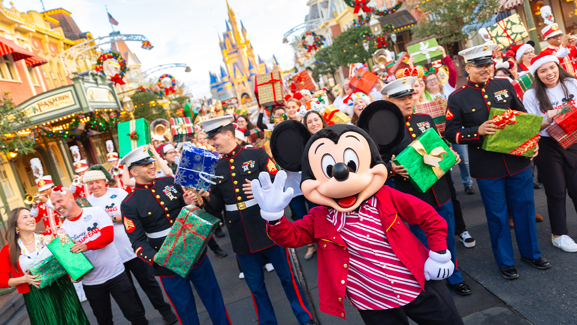 Mickey Mouse waves to the camera at Disneyland’s Main Street, U.S.A., during the Mickey Around the World Toy Drive Tour. Behind him are Disney VoluntEARS and U.S. Marines, holding wrapped gifts and smiling. The background features the Disneyland’s Sleeping Beauty Castle and shops with wreaths.