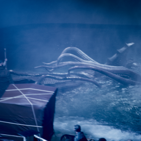 A production still depicts the filming of the incredible battle between Captain Nemo and the giant squid. Captain Nemo (played by James Mason) is standing to the left on external shell of Nautilus with cover from the highest point of the ship. He holds a spear, ready to launch it at the beast. The squid’s tentacles reach from the farthest most point of the Nautilus to Captain Nemo, eager to grab him. Its head and mouth are underwater. In the foreground of the scene is the camera, covered by a tarp, and the heads of production crew members. The entire photo has a heavy blue tint.