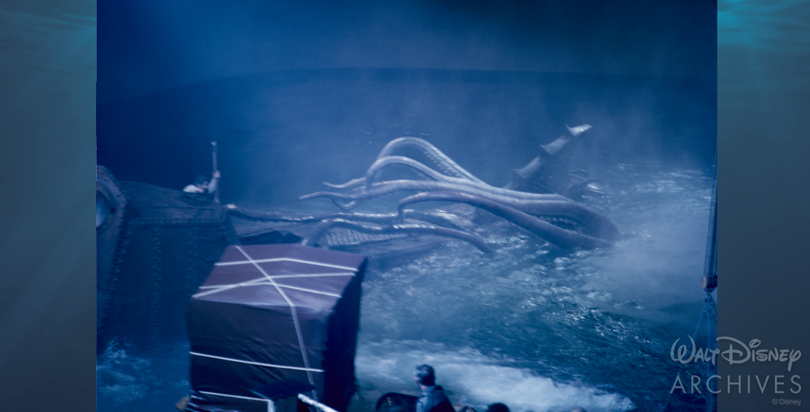 A production still depicts the filming of the incredible battle between Captain Nemo and the giant squid. Captain Nemo (played by James Mason) is standing to the left on external shell of Nautilus with cover from the highest point of the ship. He holds a spear, ready to launch it at the beast. The squid’s tentacles reach from the farthest most point of the Nautilus to Captain Nemo, eager to grab him. Its head and mouth are underwater. In the foreground of the scene is the camera, covered by a tarp, and the heads of production crew members. The entire photo has a heavy blue tint.