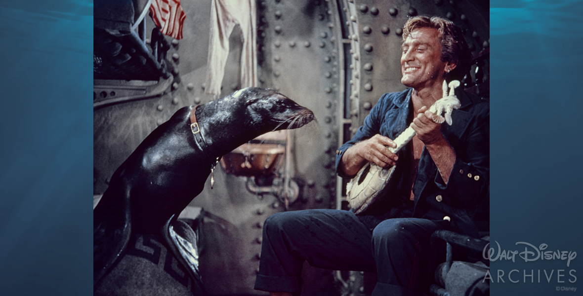 Ned Land (actor Kirk Douglas) is on the right-side bunk in a navy-colored uniform. He is holding a white guitar made from a turtle shell and seashells. On the left bunk, Esmeralda the seal sits with her face leaning toward Ned, appearing to be listening intently to the music he is playing. Esmeralda is a dark brown color with a lighter reddish-brown collar. Her flippers steady her on the gray blanket on the bed. Behind them is grayish industrial wall of Ned’s room on the Nautilus.