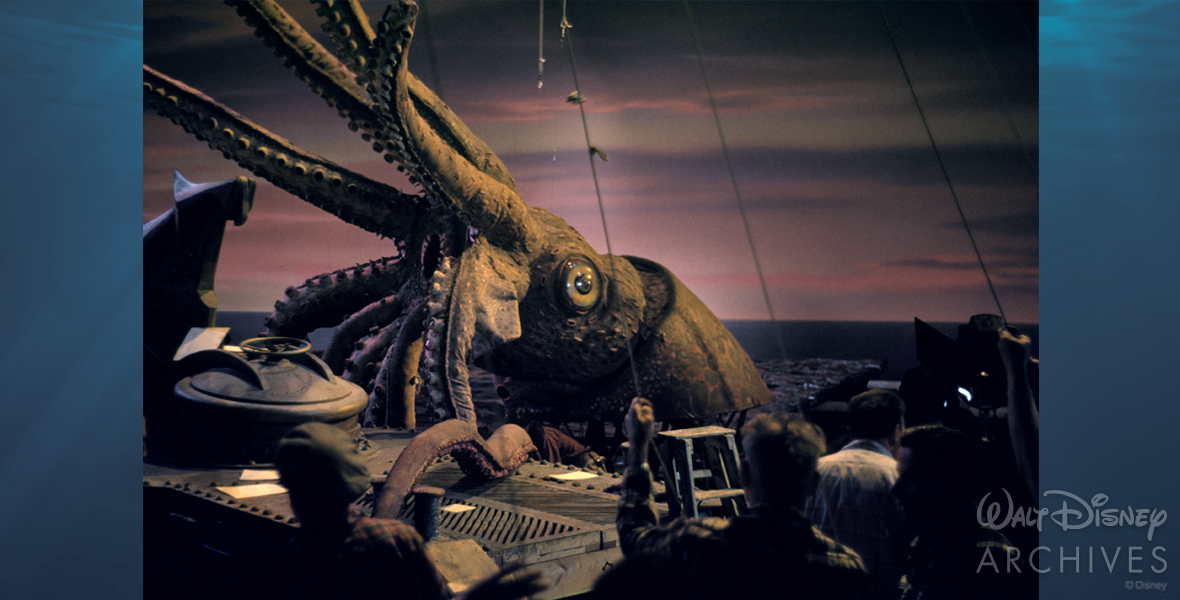 In the foreground, crew members hold on to cords that control the giant squid’s movement. The giant squid has half of its tentacles hoisted in the air and its mouth is wide open. A crew member works underneath the head. Under the head are the stilts used to hoist the squid’s head above the water in the pool of Soundstage 3 at The Walt Disney Studios in Burbank.