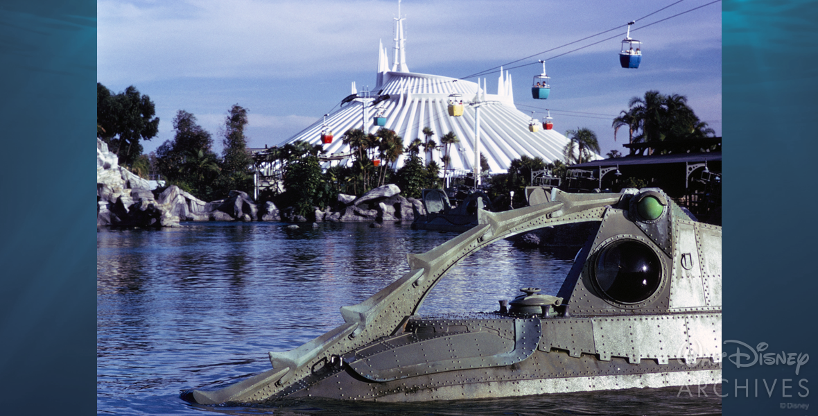 In the bottom right of the photo, the top of the Nautilus ride vehicle from the former Walt Disney World Resort attraction 20,000 Leagues Under the Sea can be seen moving through the water. One of the portholes and the spiked curved metal pieces of the ship are prominently seen. The lake is a dark blue color, expanding back to the middle of the photo, where it meets the gray rocks that mark the barrier of the attraction. In the distance the bright white Space Mountain attraction can be seen. The Skyway attraction buckets can be seen overhead, making the journey from Tomorrowland to Fantasyland, visible in between 20,000 Leagues Under the Sea and Space Mountain. They are light yellow, dark blue, light blue, and red. 