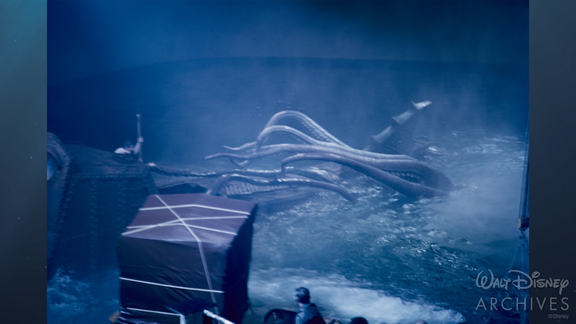 A production still depicts the filming of the incredible battle between Captain Nemo and the giant squid. Captain Nemo (played by James Mason) is standing to the left on external shell of Nautilus with cover from the highest point of the ship. He holds a spear, ready to launch it at the beast. The squid’s tentacles reach from the farthest most point of the Nautilus to Captain Nemo, eager to grab him. Its head and mouth are underwater. In the foreground of the scene is the camera, covered by a tarp, and the heads of production crew members. The entire photo has a heavy blue tint.