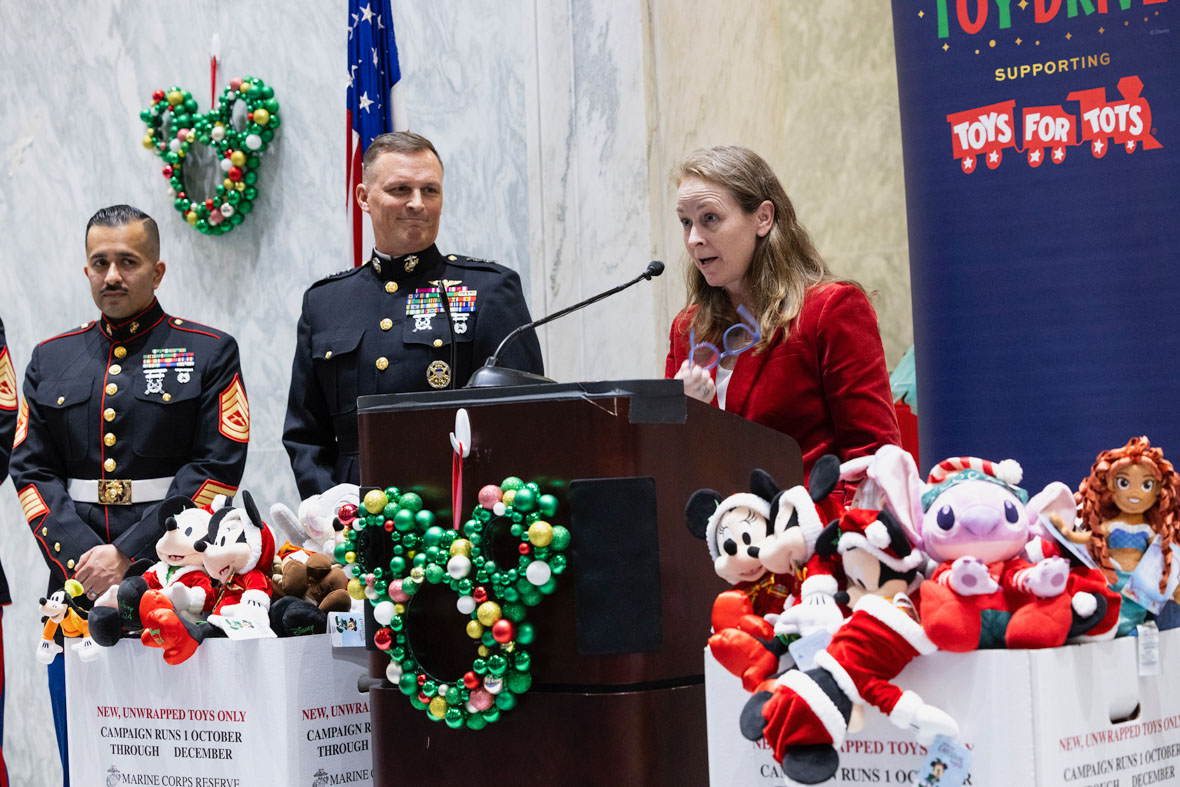 In an image from the Mickey Around the World Toy Drive Tour, a woman in a red blazer stands behind a podium and is delivering a speech. To her right are two U.S. Marines. In front of the podium are Disney plush toys.