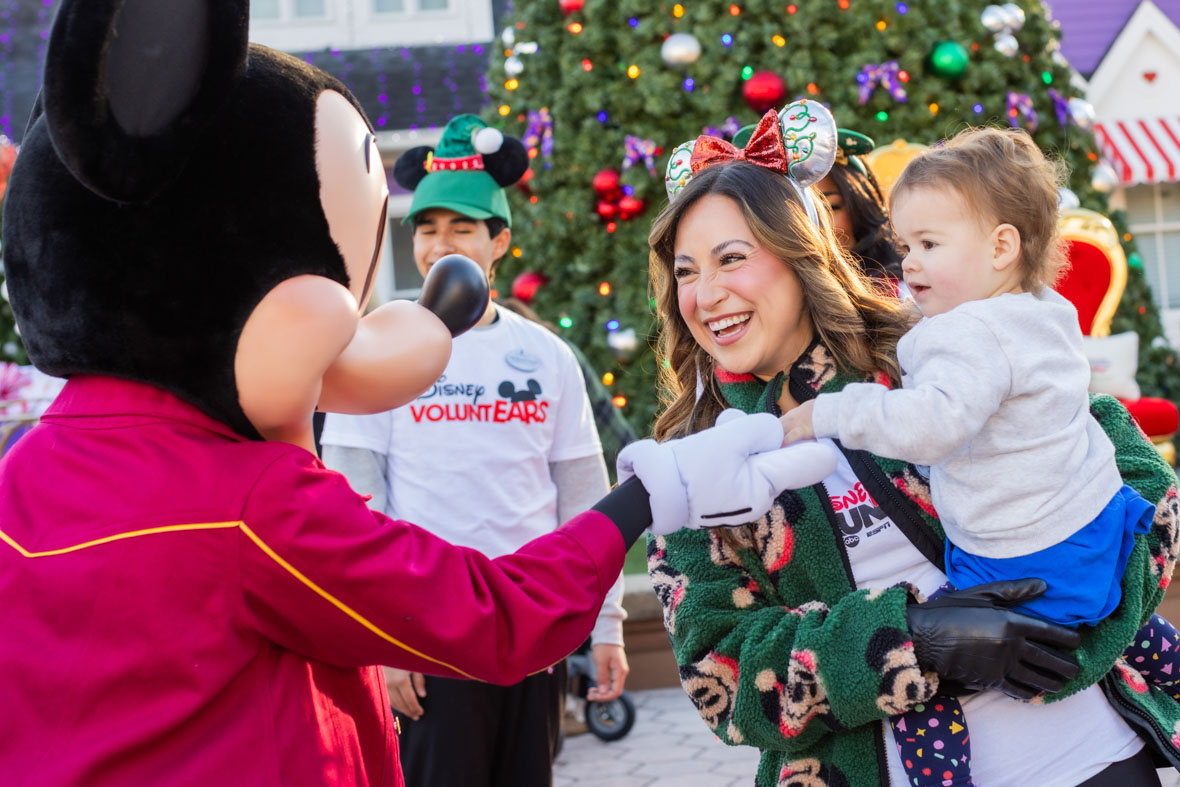 In an image from the Mickey Around the World Toy Drive Tour, Mickey Mouse interacts with a woman holding her child, smiling. The background features a lit Christmas tree and a Disney VoluntEAR.