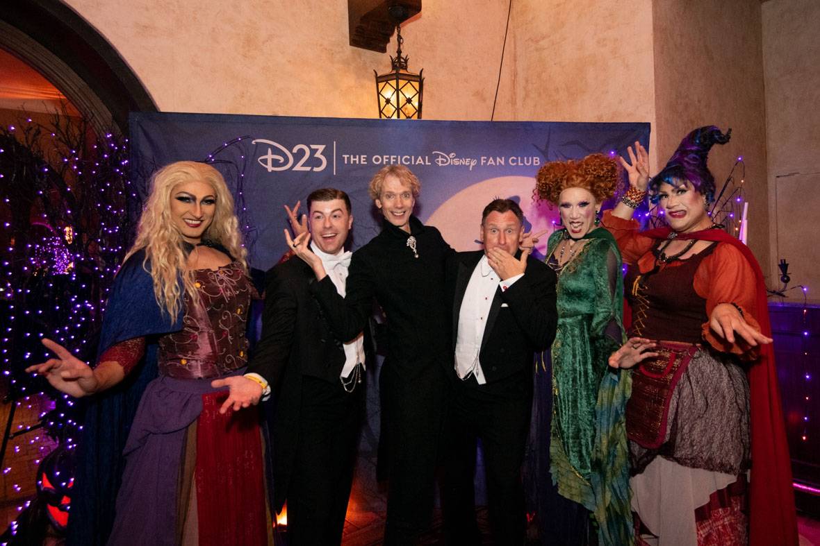 In an image from D23’s Tune-in To Terror: A Hollywood Halloween Event, Doug Jones and Sanderson Sisters performers pose with two guests in 1920’s-style suits in front of the Hocus Pocus photo op. The photo background reads “D23| THE OFFICIAL DISNEY FAN CLUB,” and there are purple Christmas lights on both sides of the frame.