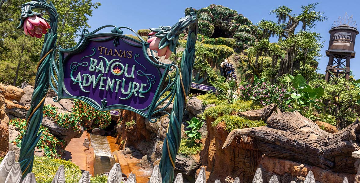 An image from outside Tiana’s Bayou Adventure attraction shows a rich purple sign with teal writing that reads: “TIANA’S BAYOU ADVENTURE.” On either side of the sign are two teal flower-shaped poles. In the background, the attraction features a mountain where guests are descending in a roller coaster car, surrounded by lush greenery.