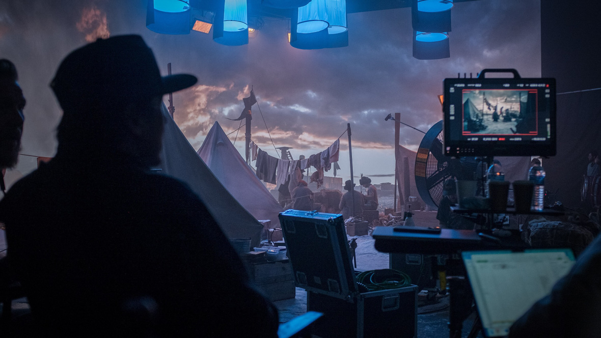 In an image from the making of National Geographic’s Endurance, director Jimmy Chin is seen in silhouette, in the foreground, on the recreation set in Los Angeles. To his right is a monitor showing an image of what his camera is seeing. In the background, a group of actors portraying Shackleton’s crew is huddled near a tent, and a backdrop of a sky is seen behind them.