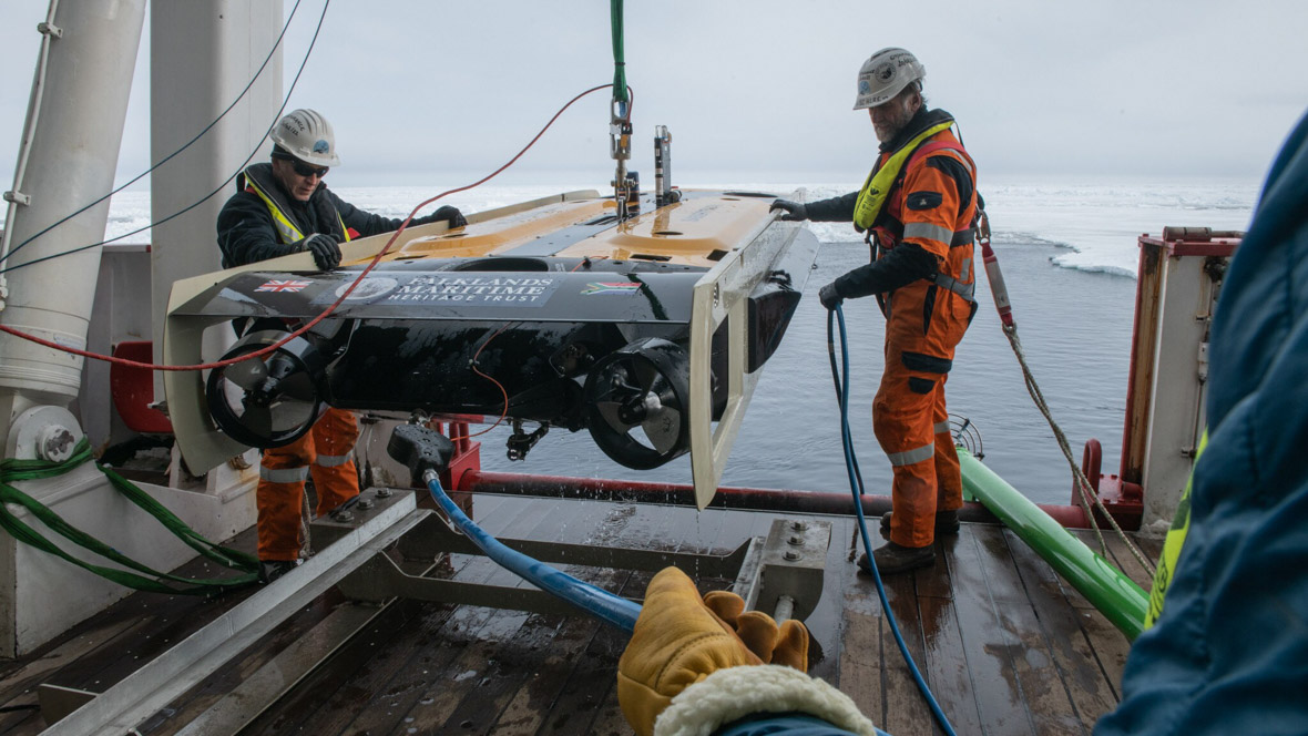 In an image from National Geographic’s Endurance, Frédéric Bassemayousse (right) and offshore manager J.C. Caillens (left) recover the AUV after a dive in search of Shackleton’s ship. Bassemayousse wears an orange jumpsuit and hard hat; Caillens wears orange pants, a dark jacket, and a hard hat. The ice-filled ocean can be seen in the background.