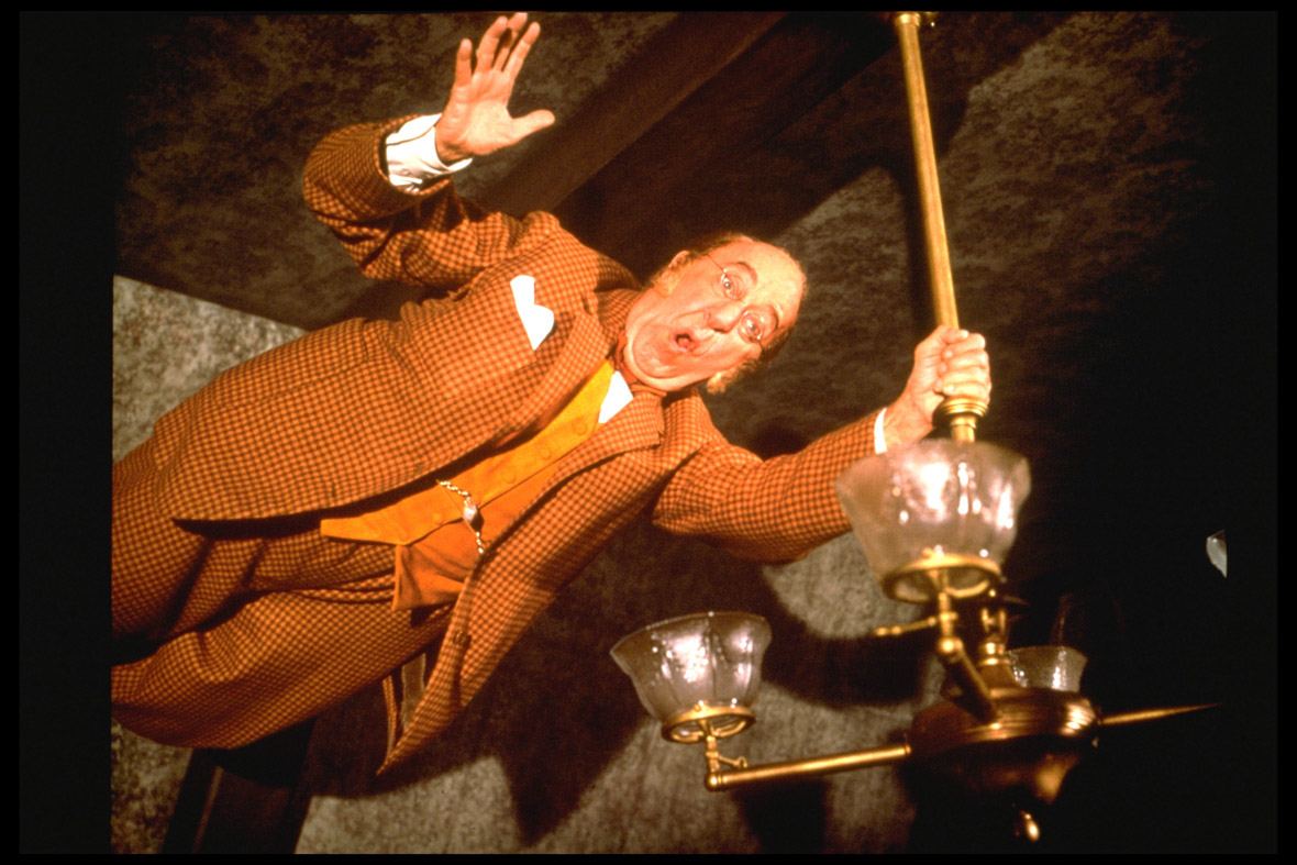 In an image from Mary Poppins, Uncle Albert (Disney Legend Ed Wynn) is holding on to a lamp as he’s floating near the ceiling while laughing uproariously. He wears a checkered orange suit and glasses.