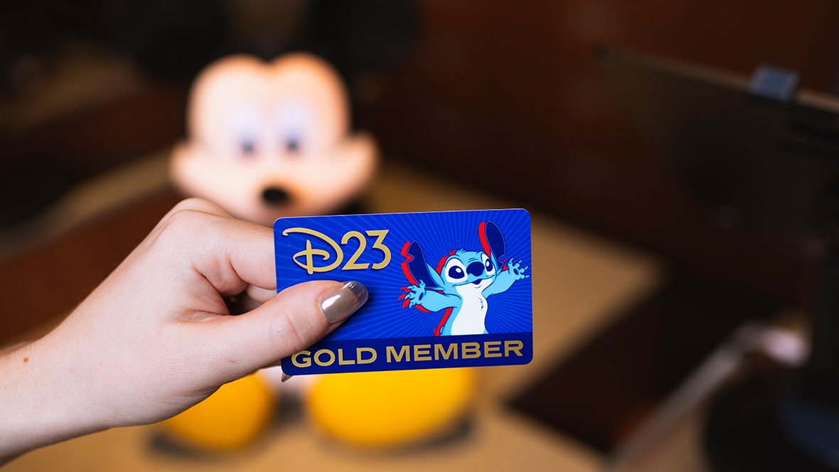 A D23 Gold Member Card being held in front of a cash register and Mickey Mouse plush.