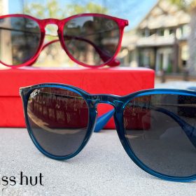 A pair of sunglasses rest on a piece of concrete. One of the sunglasses has blue frames and the other has red frames. The sunglasses with the red frames are elevated by resting on a red box. The top-right of each pair of sunglasses is adorned with the head silhouette of Mickey Mouse.