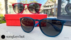 A pair of sunglasses rest on a piece of concrete. One of the sunglasses has blue frames and the other has red frames. The sunglasses with the red frames are elevated by resting on a red box. The top-right of each pair of sunglasses is adorned with the head silhouette of Mickey Mouse.