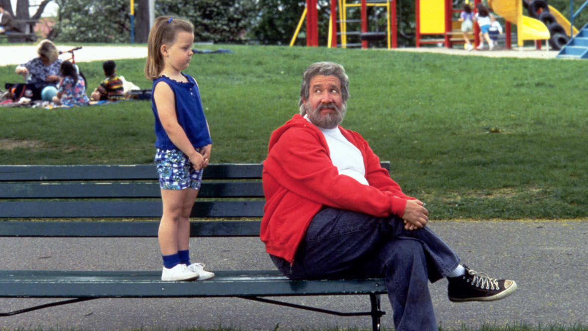 In a scene from The Santa Clause (1994), Ballet Girl (Tabitha Lupien) stands on a park bench right behind Scott Calvin (Tim Allen), who sits on the edge of the bench, looking at Ballet Girl over his shoulder. He wears a white T-shirt, a red zip-up jacket, jeans, and black converse shoes. The background features a park with grass and other children playing.