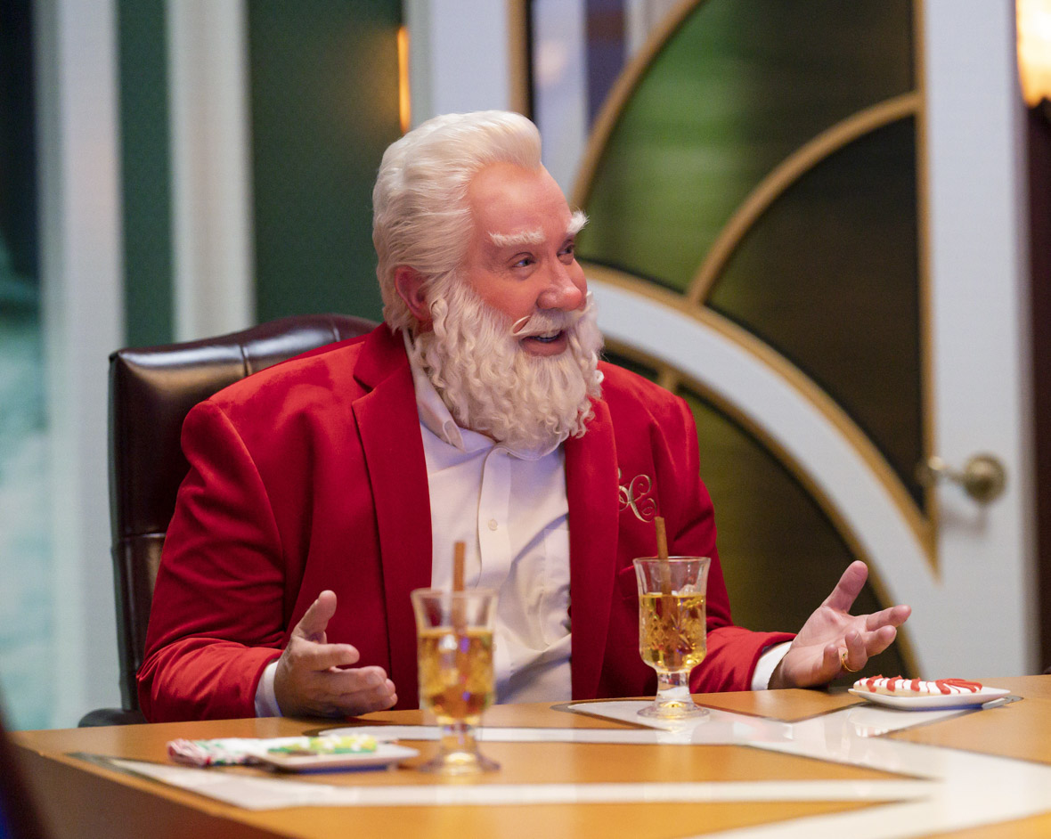 In an image from The Santa Clauses (2022), Scott Calvin (Tim Allen) is seated at a dining table, dressed in a white shirt and red jacket. He gestures with his palms facing upwards while engaging in conversation. The background shows the cozy interior of the dining room. Scott, with Santa-like signature white hair and beard, is smiling.