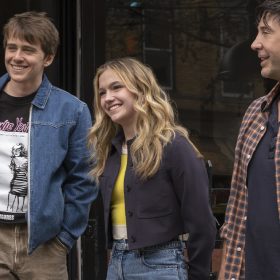 In an image from Goosebumps: The Vanishing, pictured are, from left to right, Devin (played by Sam McCarthy), Cece (played by Jayden Bartels), and Anthony (played by David Schwimmer). They are standing outside a shop. Devin and Cece look ahead, smiling, whereas Anthony looks to his right. Devin wears a black graphic T-Shirt, a denim jacket, and brown cargo pants, Cece wears a yellow top, a black crop jacket, and jeans, and Anthony wears a navy polo shirt, a flannel shirt, and jeans.