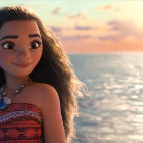 In a scene from Moana, Moana (voiced by Auliʻi Cravalho) smiles as she looks ahead. She wears a patterned strapless red top and a necklace. Her long, curly hair flows back in the wind, with the ocean and a vibrant sunset in the background.