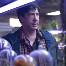 In an image from Goosebumps: The Vanishing, Anthony Brewer (David Schwimmer) stands behind shelves of bell jars containing out of focus items. He is lit with purple lighting and looking towards the right of the frame.