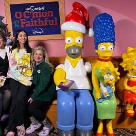 A group of four D23 guests posing with a photo opportunity for The Simpsons’ holiday special “‘O C’mon All Ye Faithful’ at the El Capitan Theatre in Hollywood. The photo opp scene features life-sized figures of Homer wearing a Santa hat and holding candy as a remote; Marge with Maggie on her lap; Lisa smiling; and Bart seated upside- down, all on their iconic living room couch. The guests are seated on the couch next to the characters, with backdrop featuring a Christmas tree, the special’s logo, and a lamp behind them, and a practical second lamp and side table to the right of the couch.