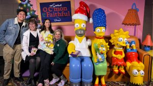 A group of four D23 guests posing with a photo opportunity for The Simpsons’ holiday special “‘O C’mon All Ye Faithful’ at the El Capitan Theatre in Hollywood. The photo opp scene features life-sized figures of Homer wearing a Santa hat and holding candy as a remote; Marge with Maggie on her lap; Lisa smiling; and Bart seated upside- down, all on their iconic living room couch. The guests are seated on the couch next to the characters, with backdrop featuring a Christmas tree, the special’s logo, and a lamp behind them, and a practical second lamp and side table to the right of the couch.