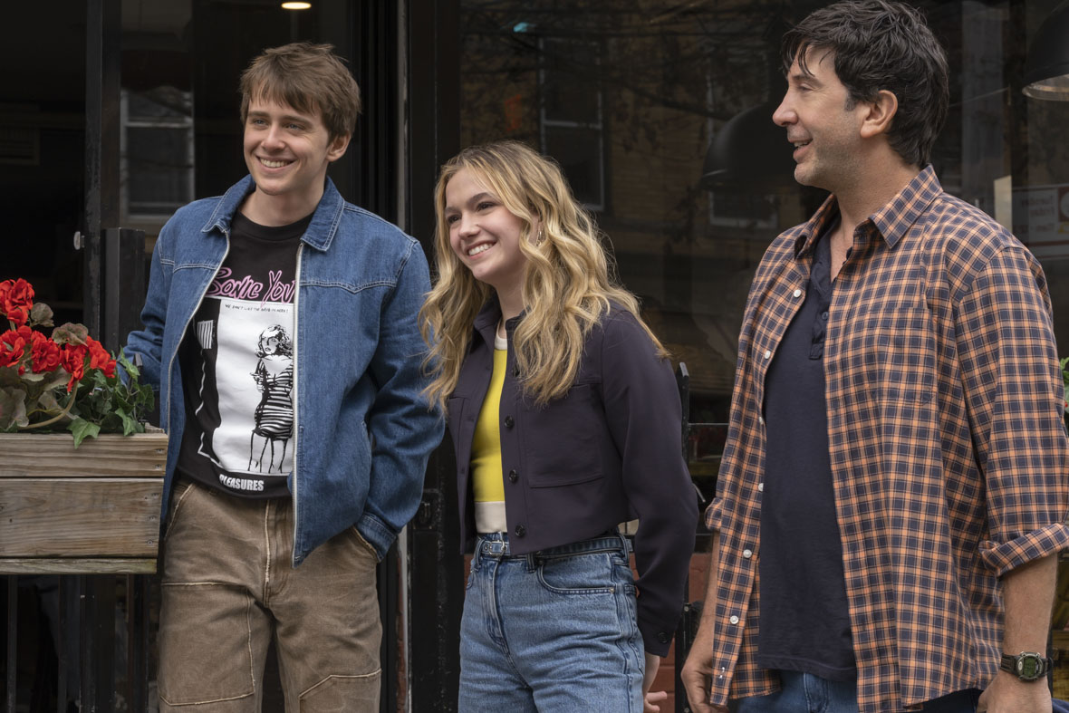 In an image from Goosebumps: The Vanishing, Devin and Cece Brewer (Sam McCarthy and Jayden Bartels) stand on the street next to their father, Anthony (David Schwimmer), in front of a window. Devin and Cece are looking ahead and smiling, while Anthony is smiling at his two children. 